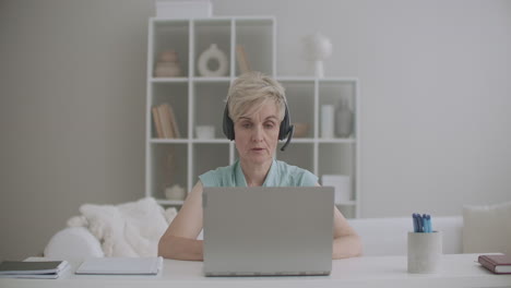 aged-woman-specialist-is-consulting-online-working-from-home-using-laptop-with-internet-and-headphones-indoors-shot